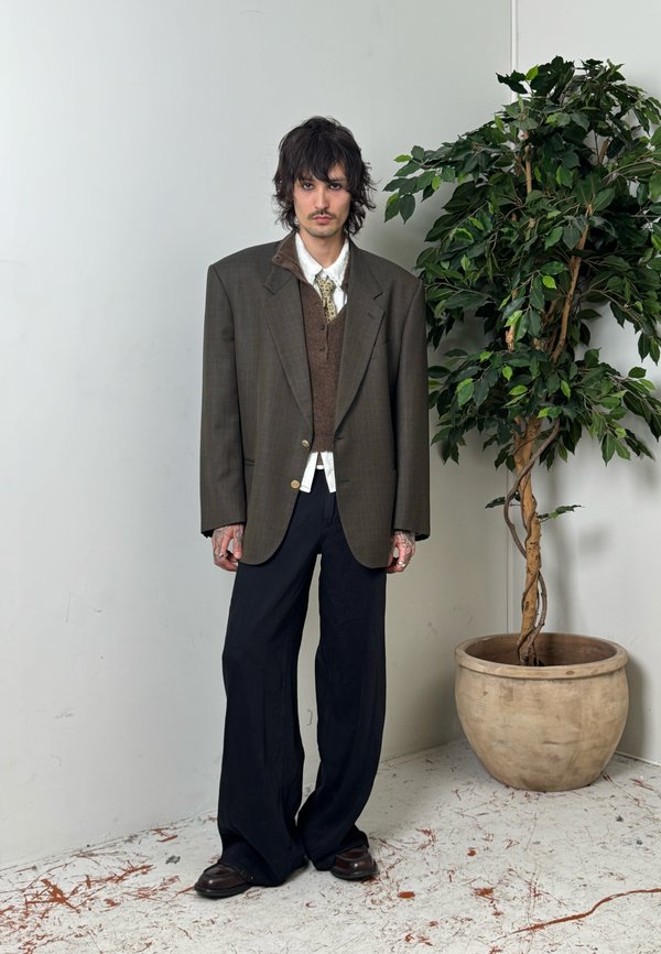 Hombre con cabello oscuro de longitud media, vistiendo una sobredimensionada chaqueta marrón, camisa blanca, corbata estampada y pantalones anchos oscuros, de pie junto a una gran planta frondosa en maceta.
