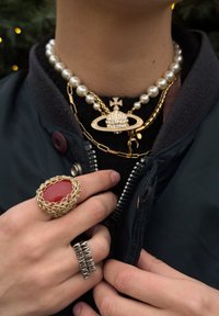Personne portant des colliers superposés en or et perles avec un pendentif croix orné de bijoux, ainsi que deux bagues, l'une avec une pierre rouge et l'autre avec un texte.