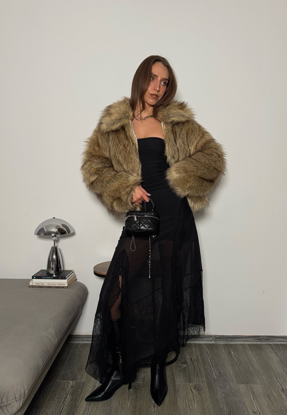 Veste en fausse fourrure beige, robe bustier noire avec accents de dentelle, et bottines noires pointues. Petit sac noir matelassé à la main.