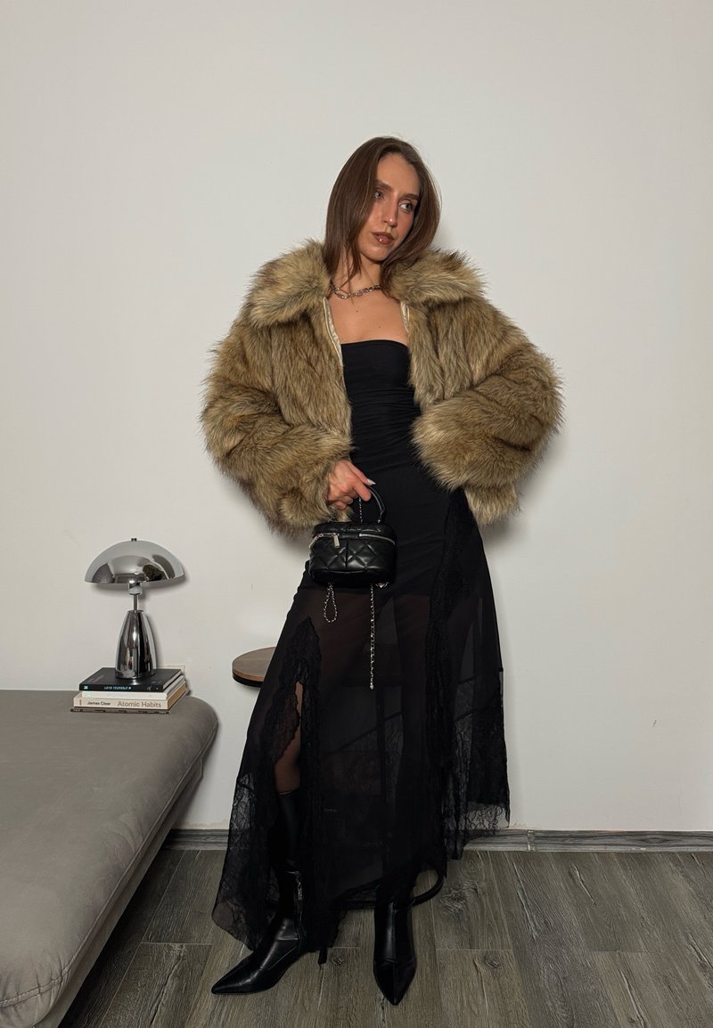 Veste en fausse fourrure beige, robe bustier noire avec accents de dentelle, et bottines noires pointues. Petit sac noir matelassé à la main.