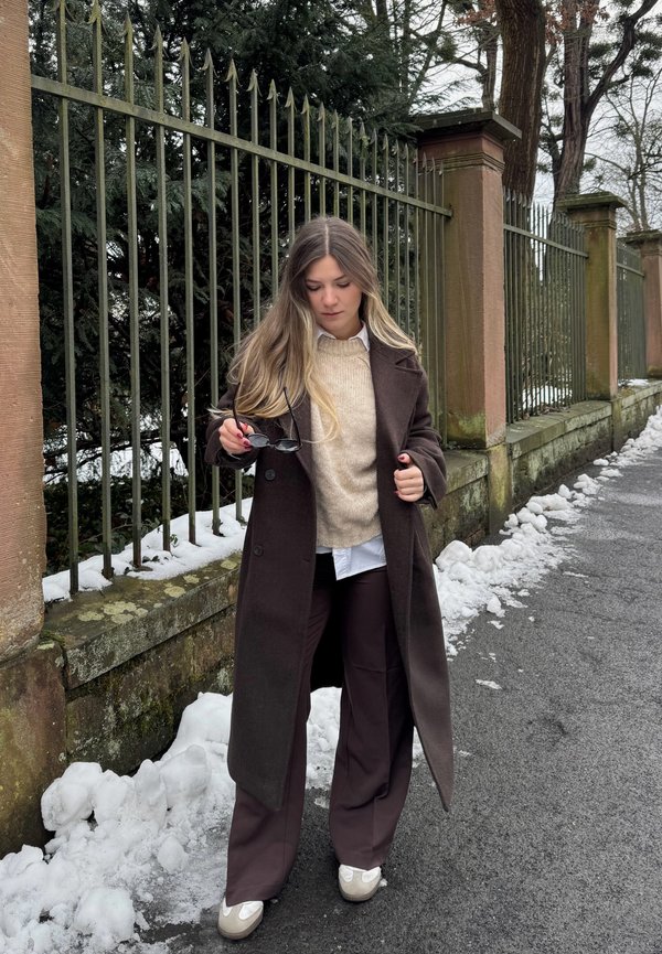 Žena v hnědém kabátu a kalhotách držící sluneční brýle, stojící na zasněženém chodníku vedle železného plotu a kamenných sloupů s stromy v pozadí.