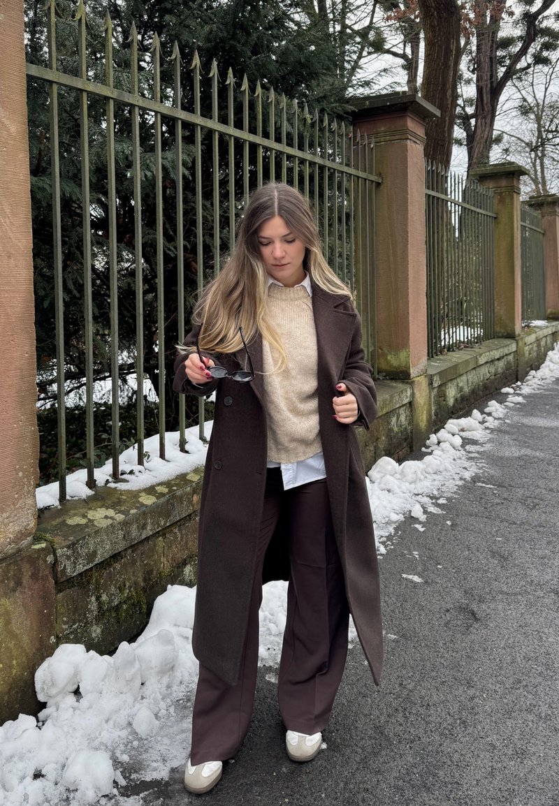 Femme en manteau et pantalon marron tenant des lunettes de soleil, debout sur un trottoir enneigé à côté d'une clôture en fer et de piliers en pierre avec des arbres en arrière-plan.