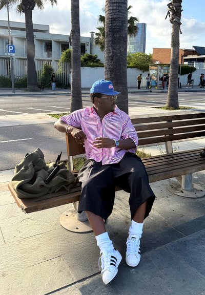 Camisa a rayas rojas y blancas, pantalones cortos negros, zapatillas blancas con rayas negras y una gorra azul. Sentado en un banco de madera al aire libre.