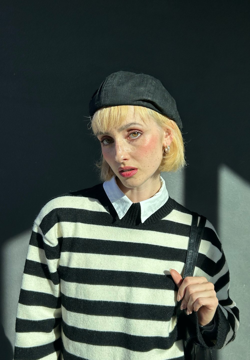 Ριγέ πουλόβερ σε μαύρο και κρεμ χρώμα με V-λαιμό, σε συνδυασμό με λευκή πουκαμίσα με κολάρο. Μαύρο μπερέ και ασημένιες κρίκοι αυτιών ολοκληρώνουν την εμφάνιση.