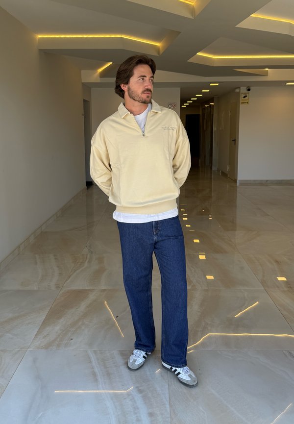 Hombre con suéter beige, camisa blanca, jeans azules y zapatillas de deporte de pie en un pasillo moderno con suelo de mármol reflectante y luces empotradas en el techo.