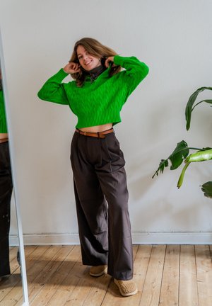 Joven sonriente ajustándose el cabello, vistiendo un suéter corto verde brillante, pantalones anchos marrones y pantuflas beige, de pie en interiores junto a una planta.