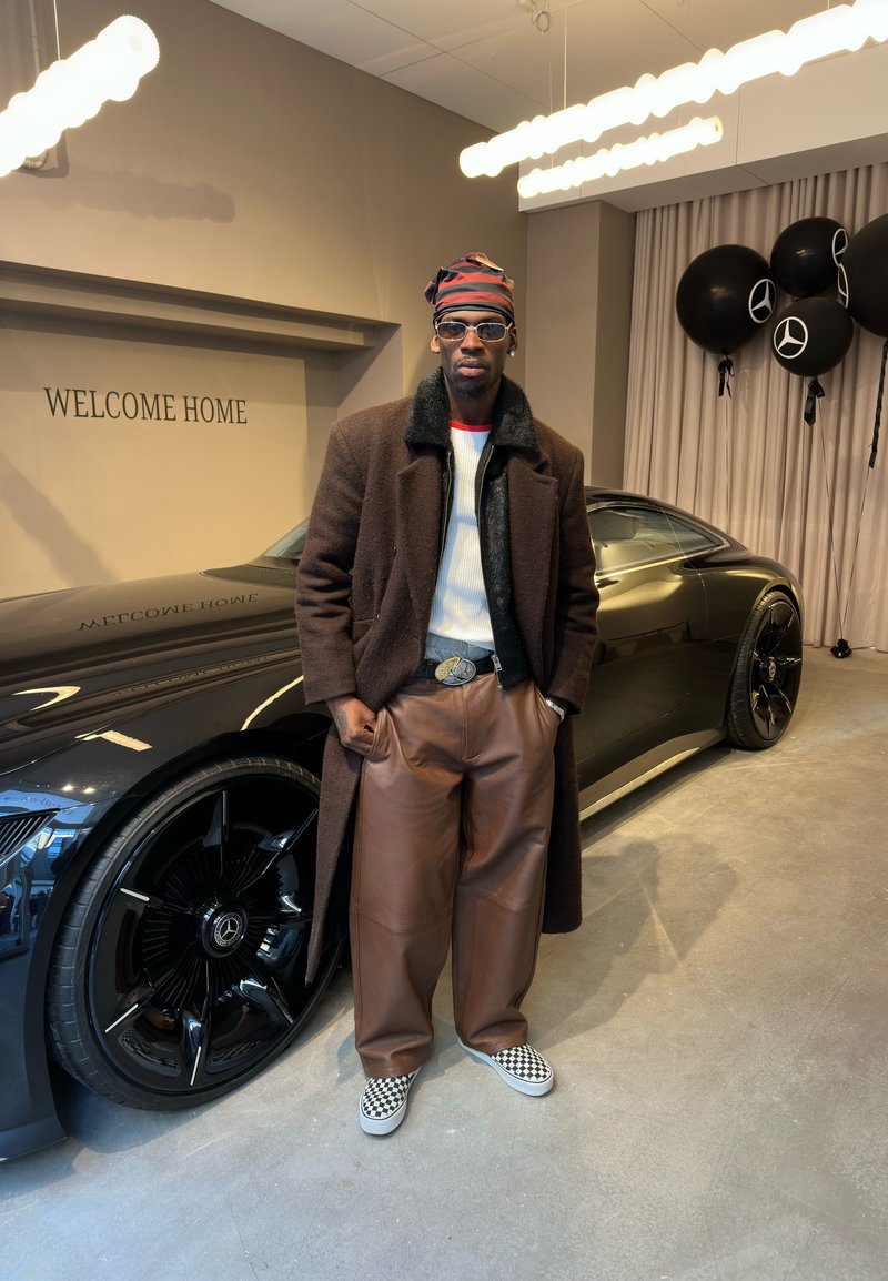 Homme portant un manteau marron, un pantalon en cuir et des chaussures slip-on à carreaux, debout à côté d'une voiture de luxe noire avec des logos Mercedes et des ballons noirs.