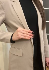 Femme portant un blazer beige et un haut noir, montrant des ongles manucurés foncés avec la main posée sur le devant du blazer.