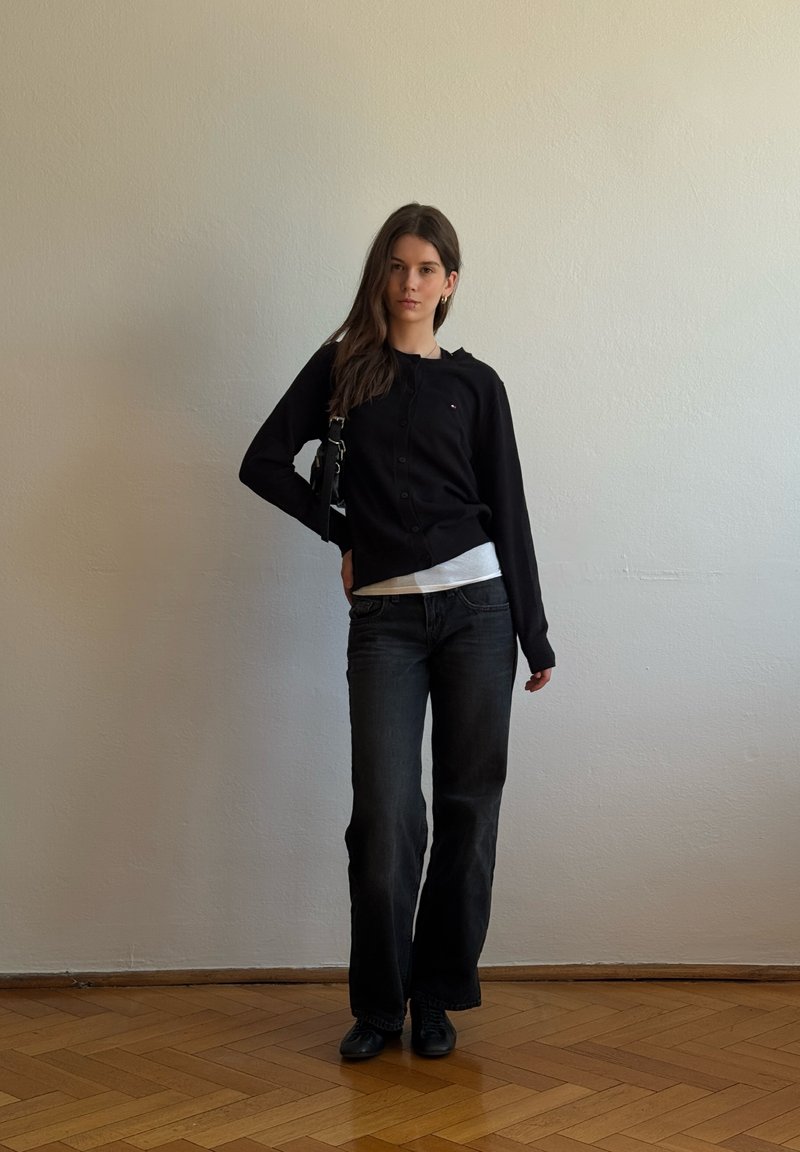 Jeune femme debout à l'intérieur portant un gilet noir, un jean foncé, des chaussures noires, et portant un petit sac à bandoulière noir devant un mur blanc uni.