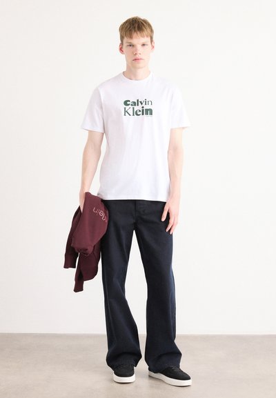 Camiseta blanca con el logo verde de "Calvin Klein", pantalones vaqueros de pierna ancha de color azul oscuro y un sudadera color marrón caoba colgando del brazo. Zapatillas negras en los pies.