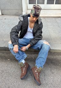 Črni usnjeni bomber jakna z umetnim krznom, bela majica brez rokavov, raztrgane modre kavbojke, rjavi pohodniški čevlji z zelenimi poudarki in kamuflažna kapa beanie.