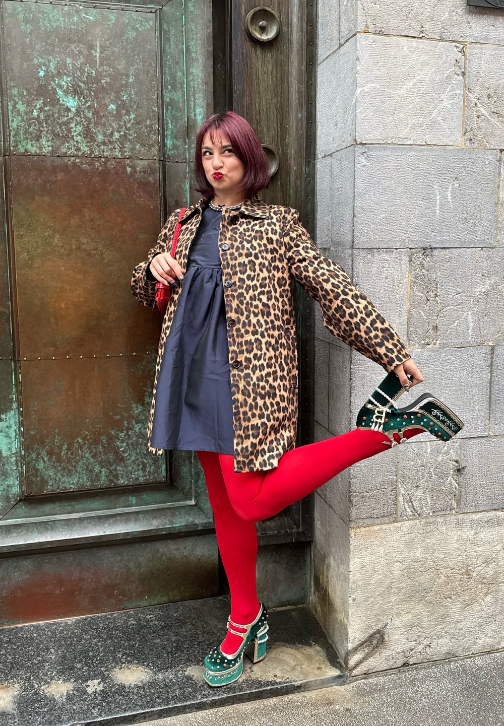 Leopardenmuster Mantel, marineblaues Kleid, leuchtend rote Strumpfhosen, grüne Plateauschuhe mit Nieten und einem Riemen. Steht neben einer strukturierten Metalldoor.
