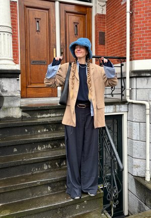 Blazer beige sobre un suéter azul marino, pantalones anchos de rayas en azul marino, y un sombrero tipo cubo azul, con un bolso negro colgado del hombro.