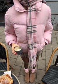 Lyserød puffedjakke, ternet tørklæde med lyserøde og grå detaljer, holder en mørk kop. Lysebrune UGG-støvler og et ternet skørt er synlige.