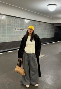 Woman wearing a yellow beanie, glasses, a white blouse, a long black coat, and wide grey trousers, holding a tan handbag, standing in Schönleinstraße subway station.
