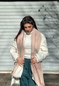 Ljusrosa och beige rutig halsduk med fransiga kanter, bärs över en vit tröja och en lång vit kappa, kombinerad med blågröna byxor.