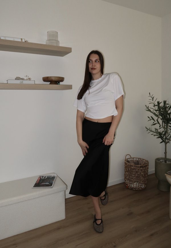 Kvinde i hvid cropped t-shirt og sort nederdel står indendørs nær hylder, magasin, flettet kurv og potteplante på trægulv.