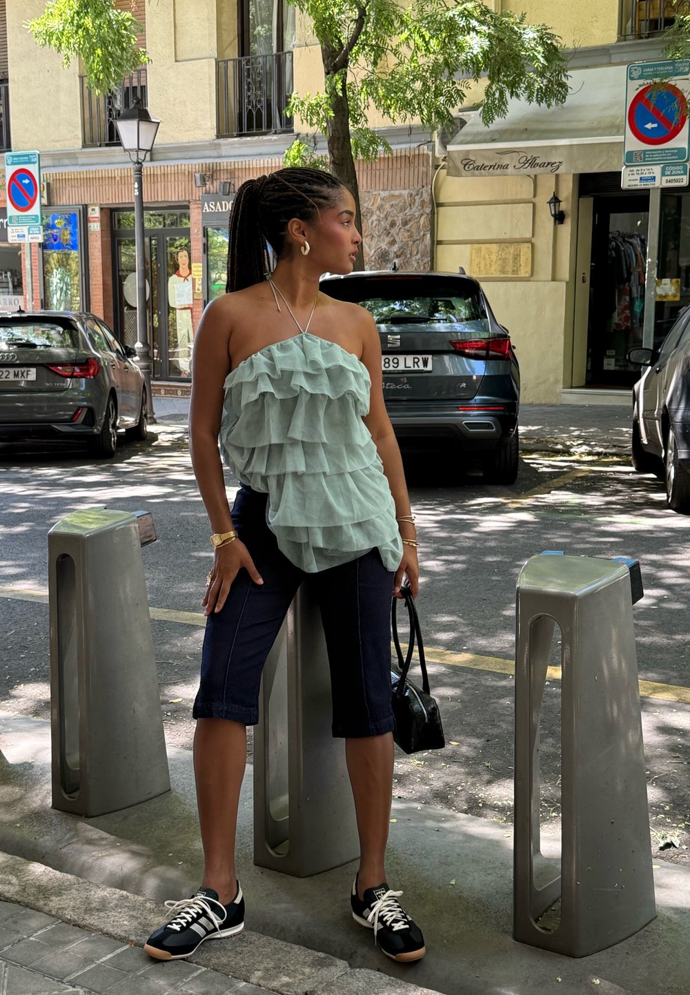 Jeune femme aux cheveux tressés portant un haut vert menthe à volants et un pantalon capri noir, debout dans une rue ensoleillée de la ville entre des voitures garées.