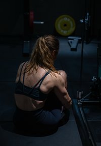 Athletic woman with a ponytail stretching while seated on the gym floor, near weightlifting equipment in a dimly lit workout space.