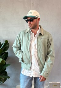 Hele rohelise jope beeži kraega, kantud valge nööbitava särgi kohal. Mustad päikeseprillid ja valge-roheline müts. Taustal taimed.