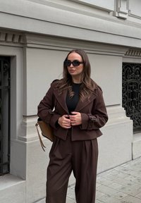 Καφέ σακάκι από μίγμα μάλλινου υλικού με μανίκια που έχουν ζώνη, συνδυασμένο με μαύρη μπλούζα και φαρδιά καφέ παντελόνια. Συνοδεύεται από μαύρα γυαλιά ηλίου και μια καφέ τσάντα.