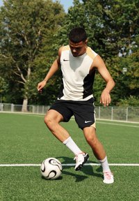 Fotballspiller i en ermeløs hvit og svart drakt med gullakenter, svarte shorts og hvite strømper, som dribler en svart-hvit fotball på kunstgress.