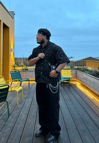 Chemise noire à boutons avec des clous métalliques, pantalon noir ample, accents en chaîne argentée et béret noir. En arrière-plan, on trouve une terrasse en bois et des chaises jaunes.