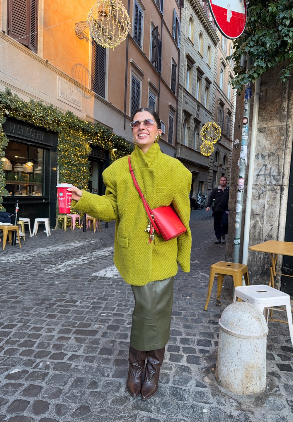 Manteau vert vif surdimensionné, jupe vert olive, bottes hautes marron, sac bandoulière rouge. Rue pavée avec décorations festives en arrière-plan.