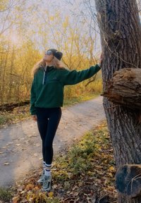 Zielony polar z półzamkiem i kieszenią na piersi, w połączeniu z czarnymi legginsami i białymi skarpetkami w paski, na tle naturalnej ścieżki na zewnątrz.