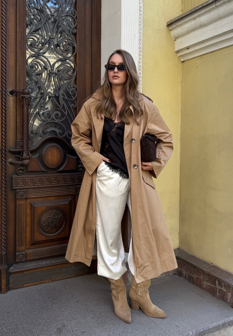 Trench coat beige su un top in pizzo nero, pantaloni in satin crema e stivaletti alla caviglia in suede beige. Gli accessori includono occhiali da sole neri e una clutch marrone.