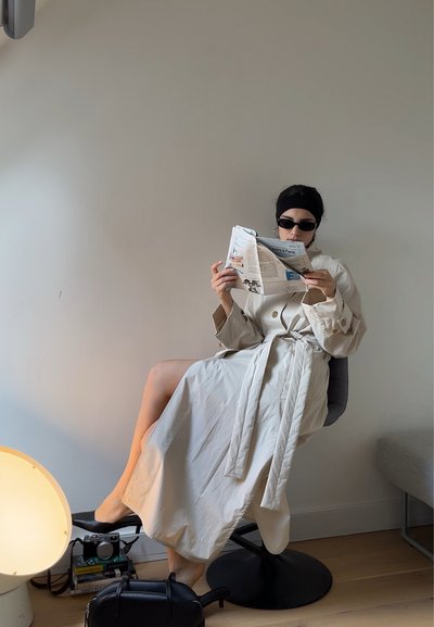 Larga gabardina beige con un cinturón en la cintura, llevada sobre una blusa ajustada. La persona está sentada, leyendo un periódico mientras lleva gafas de sol.