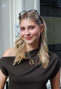 Brauner, schulterfreier Oberteil, goldene, runde Anhängerhalskette, goldene Creolen, lange blonde Haare, dezentes Make-up und eine Brille, die auf ihrem Kopf ruht.