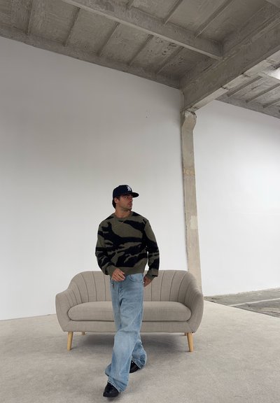 Hombre vestido con suéter de camuflaje oscuro, jeans azul claro holgados, zapatos negros y gorra de béisbol oscura, de pie frente a un sofá beige en una habitación minimalista.