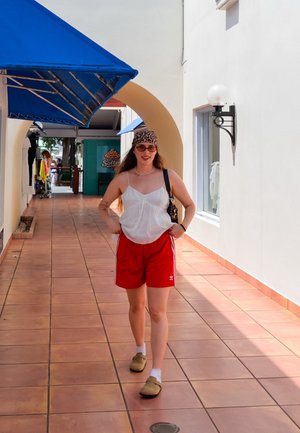 Top sin mangas blanco, pantalones cortos rojos con rayas blancas, zapatos de charol color beige, y una diadema de estampado de leopardo, caminando por un corredor de azulejos con toldos azules.