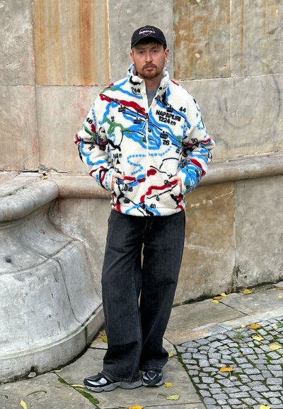 Chaqueta de forro polar con estampado de mapa en color crema, que presenta acentos en azul, rojo y verde. Combinada con pantalones anchos oscuros y zapatillas grises.