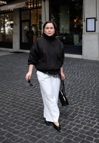 Nainen, jolla on päällä musta takki, valkoiset housut ja mustat korkokengät, pitää kädessään aurinkolaseja ja mustaa käsilaukkua, kävelee mukulakivikadulla liikkeen edustalla.