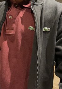 Jachetă neagră cu fermoar peste un tricou polo bordeaux. Polo-ul are un material texturat și un mic logo de crocodil verde pe piept.