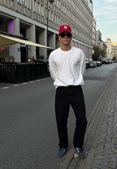 Una persona está de pie en una calle, vistiendo una camisa de manga larga blanca texturizada, pantalones negros, una gorra roja y zapatillas gris. El entorno es urbano.