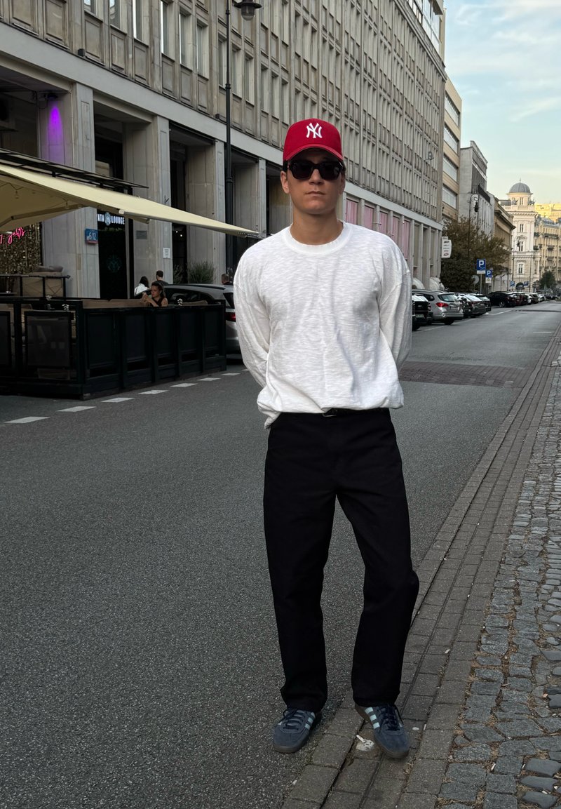Una persona si trova in una strada indossando una camicia bianca a maniche lunghe con texture, pantaloni neri, un cappellino rosso e sneakers grigie. L'ambientazione è urbana.