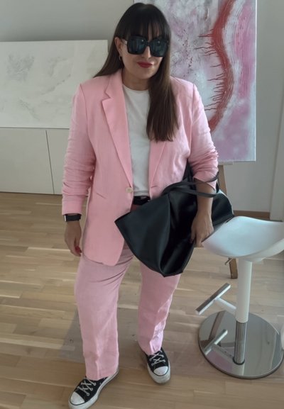 Traje de lino rosa con un blazer ajustado y pantalones de pierna ancha, combinado con una camiseta blanca y un bolso de mano negro. Zapatillas negras completan el look.