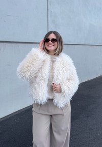 Νεαρή γυναίκα που φοράει μια αφράτη λευκή ζακέτα, μπεζ παντελόνια, γυαλιά ηλίου, χαμογελώντας, στέκεται μπροστά σε έναν γκρι τσιμεντένιο τοίχο πάνω σε άσφαλτο.