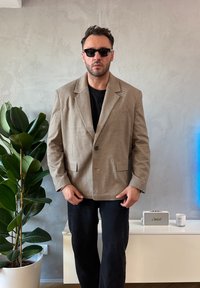 Barna és bézs vékony csíkos blézer két első zsebbel, fekete ing fölött viselve, fekete nadrággal párosítva. A háttér zöld növényzetet ábrázol.