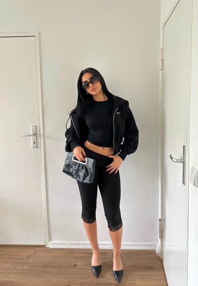Mujer con top corto negro, chaqueta, leggings con encaje, tacones y gafas de sol, sosteniendo un bolso negro, de pie en el interior junto a dos puertas blancas.