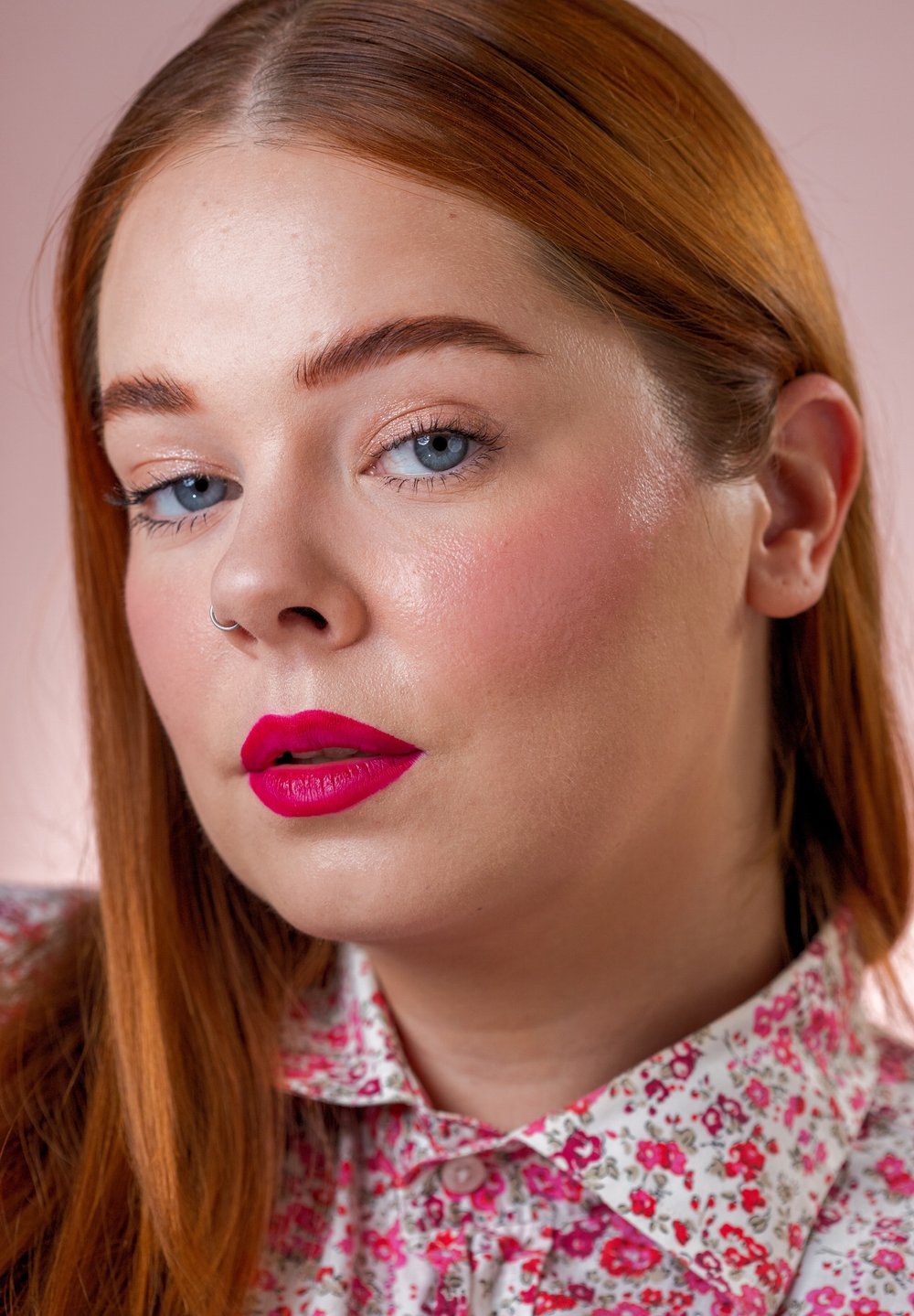 Close-up van een persoon met lang, steil, rood haar. Ze hebben felle roze lippenstift, blauwe ogen en dragen een bloempatronen shirt.