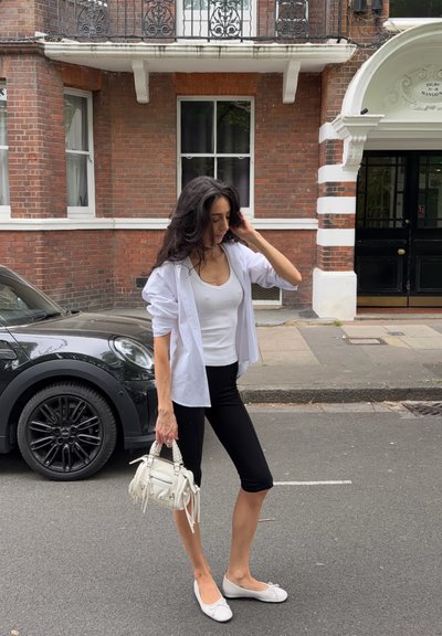 Mujer con camisa blanca, camiseta sin mangas y leggings negros, sosteniendo un bolso blanco, de pie en una calle con un coche negro y un edificio de ladrillos detrás.