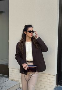 Γυναίκα με μακριά σγουρά μαλλιά που φοράει μαύρα γυαλιά ηλίου, σκούρο σακάκι, λευκό πουλόβερ, μπεζ παντελόνι και κρατάει μια μαύρη τσάντα φάκελο δίπλα σε ένα μπεζ τοίχο.