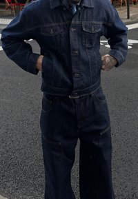 Mörk tvättad jeansjacka och byxor med synlig sömnad, knappstängningar och framfickor. Löst snitt med en något överdimensionerad stil.