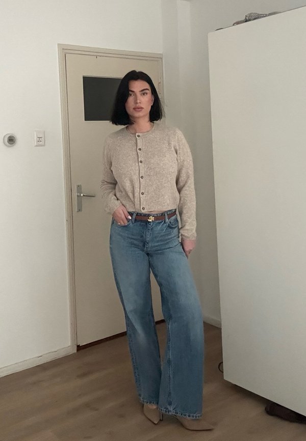 Woman with black bob hair wearing beige buttoned cardigan, wide-leg blue jeans, brown belt, and beige pointed shoes standing indoors.