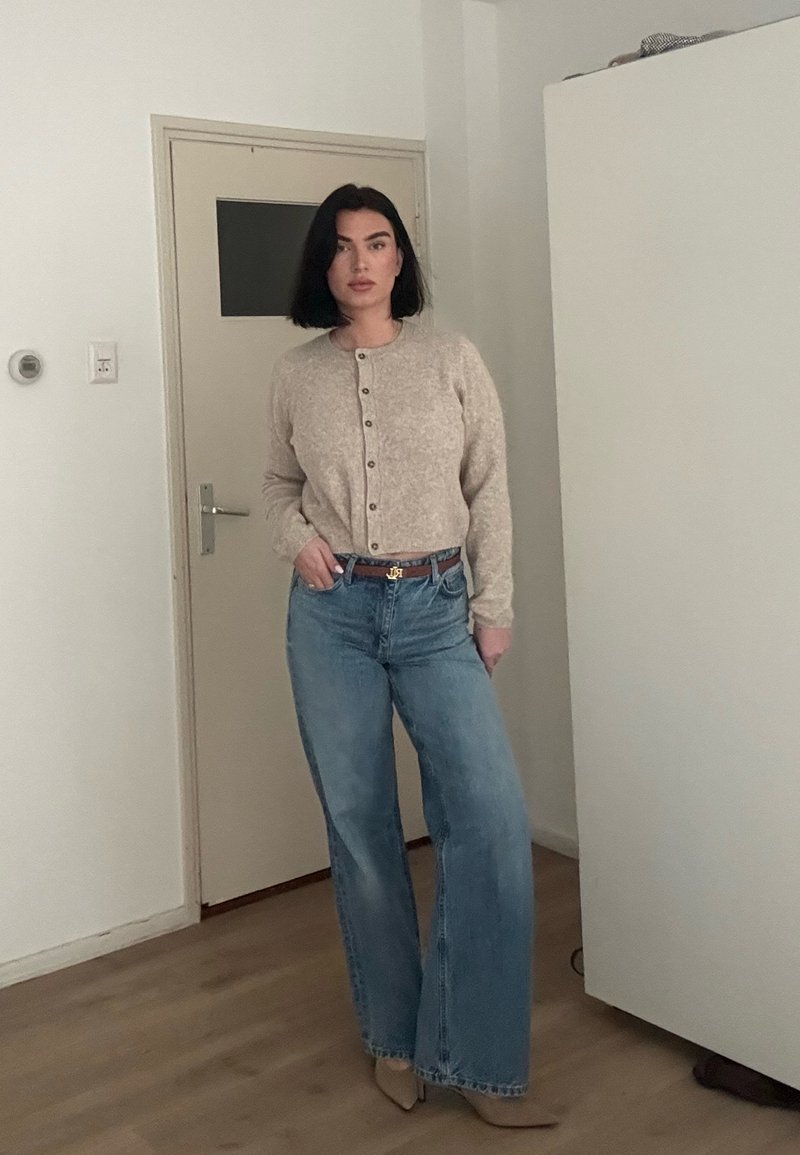 Femme aux cheveux noirs en coupe au carré, portant un cardigan beige boutonné, un jean bleu large, une ceinture marron et des chaussures beiges à bout pointu, debout à l'intérieur.