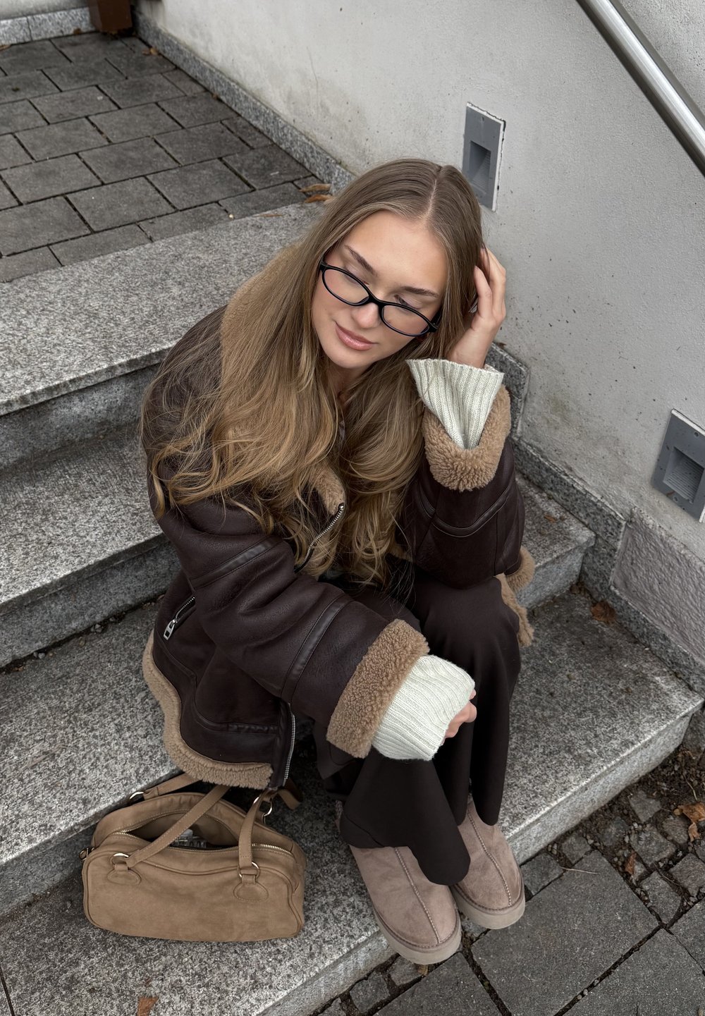 Jachetă maro din blană ecologică cu margini crem, asortată cu pantaloni maro închis. Geantă bej de tip suede și cizme de gleznă asortate. Fundal cu trepte din piatră gri.